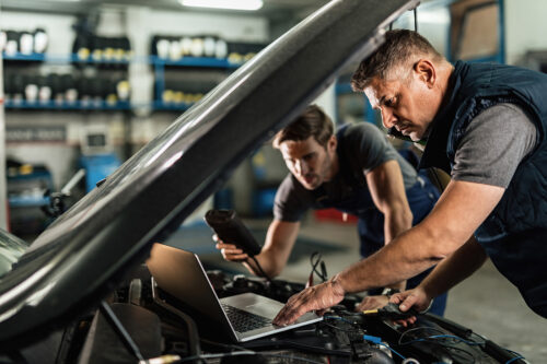Auto mechanic using laptop and cooperating with a coworker while doing car diagnostic in repair shop.